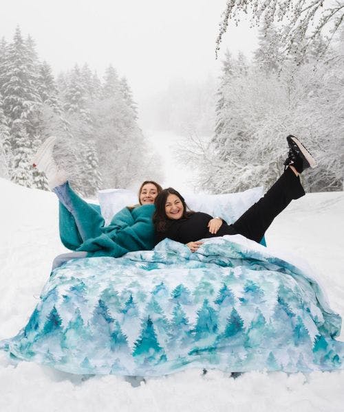 Deux femmes allongées sur un lit en extérieur dans la neige. Le lit porte une parure de lit fabriquée en France par Blanc de Gérardmer.