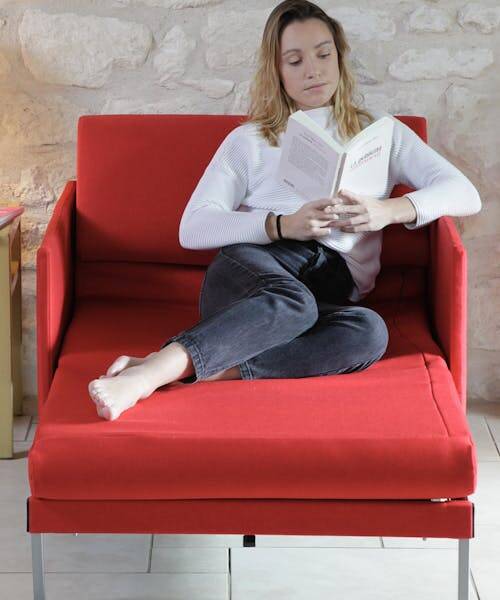 Femme blonde entrain de lire un livre assise sur un fauteuil rouge convertible et fabriqué en France de la marque Likoolis.
