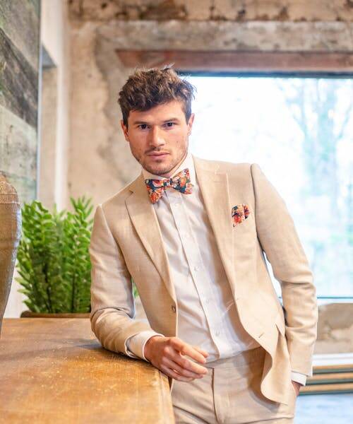 Homme châtain portant un costume beige accompagné d'une pochette de costume et d'un noeux papillon liberty fabriqués en France par Gentille Alouette.