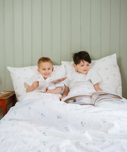 Deux garçons, portant des pyjamas Made in France, lisent un livre dans un lit équipé d'une parure fabriquée en France par Cotonboul.